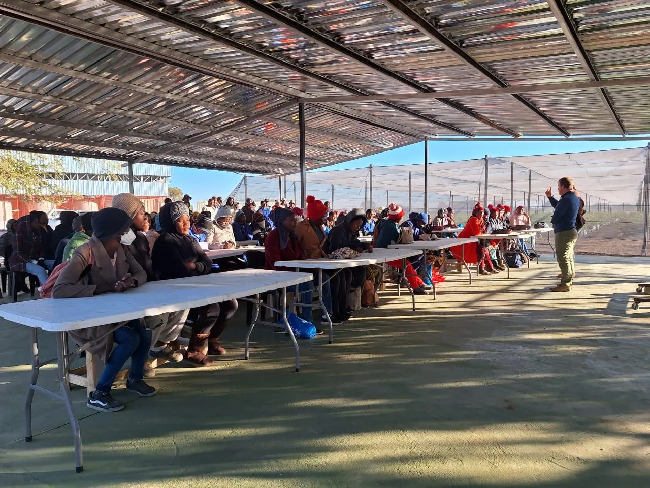 Community members seated at long tables under a shade roof during a meeting on the farm