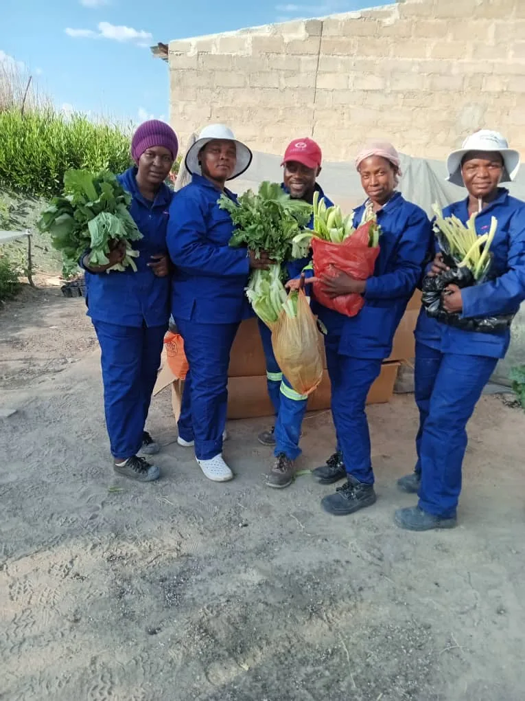 Five women in blue overalls holding freshly harvested vegetables